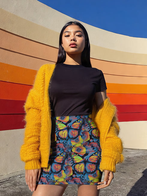 High-waisted bodycon mini skirt with an allover print of rainbow monarch butterflies and colorful tropical flowers on a dark background. Soft, stretchy poly-spandex fabric hugs the body for a flattering fit. Styled for rave, Y2K, and festival outfits. Sizes XS to XL.